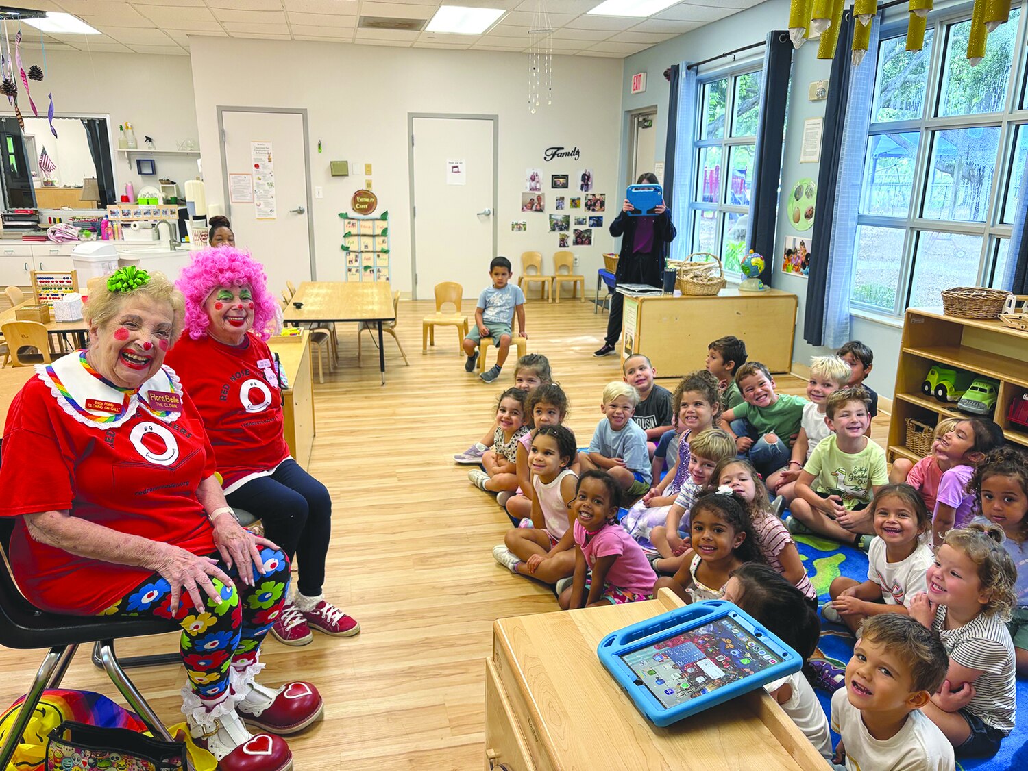 Two clowns seated with a large group of preschoolers in a bright classroom
