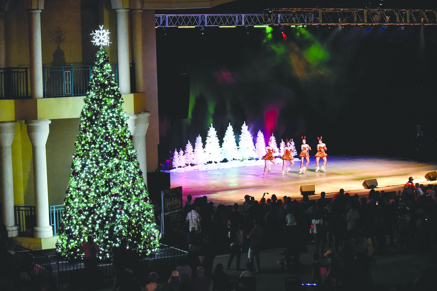 Lighted holiday tree beside skaters performing on an outdoor stage
